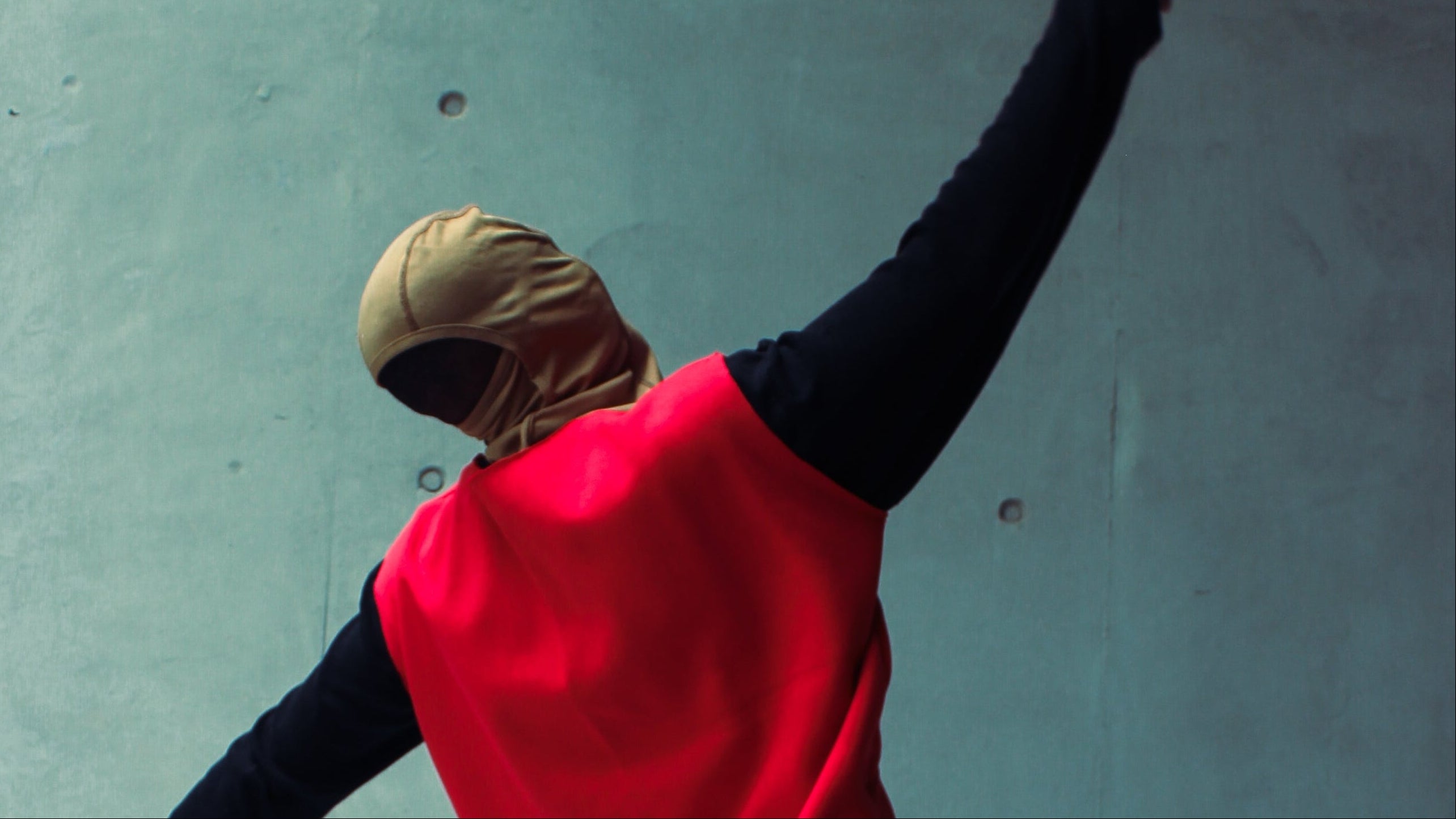 Person in red vest and black pants standing on a white mat with arms outstretched against a concrete wall.