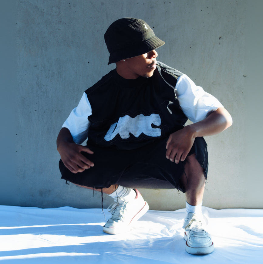 Person wearing a black vest with a logo, white shirt, and sneakers against a concrete wall.