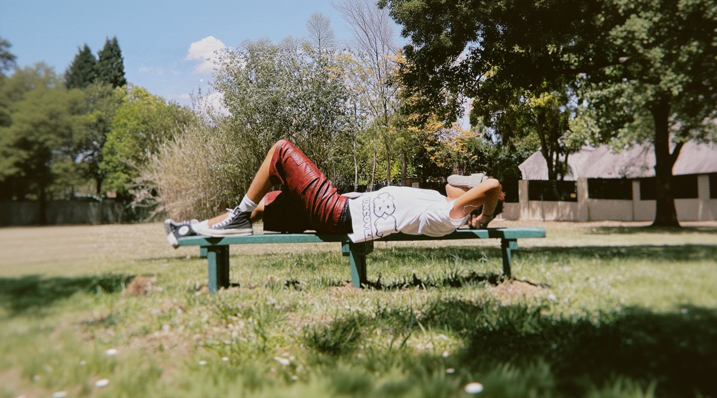 Person lying on a bench in a park with trees and grass
